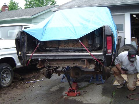 1979 Ford Bronco - A man works on the rear axle of a disassembled truck, which is partially covered by a blue tarp.