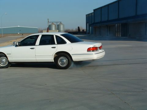 1995 Ford Crown Victoria - A white 1995 Ford Crown Victoria is captured mid-burnout on a paved lot.