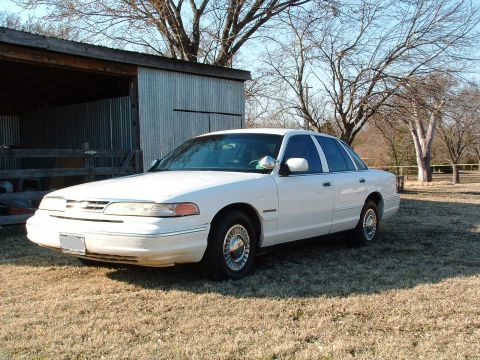 1995 Ford Crown Victoria - A white 1995 Ford Crown Victoria is parked on a grassy area near a rustic shed.