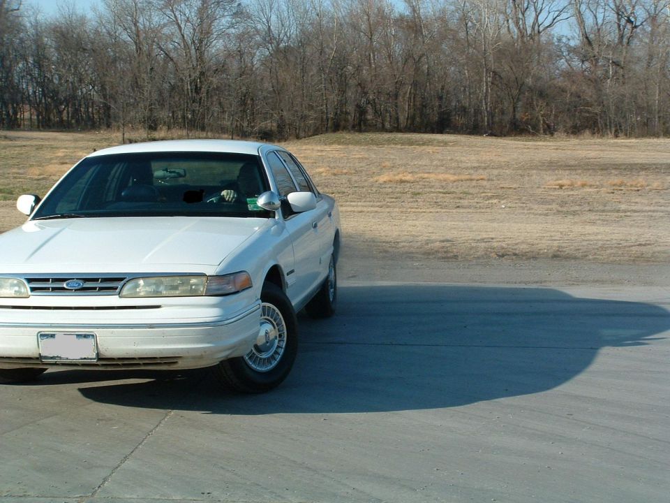 White 1990s Ford Crown Victoria sedan parked on concrete pad with bare trees and field behind
