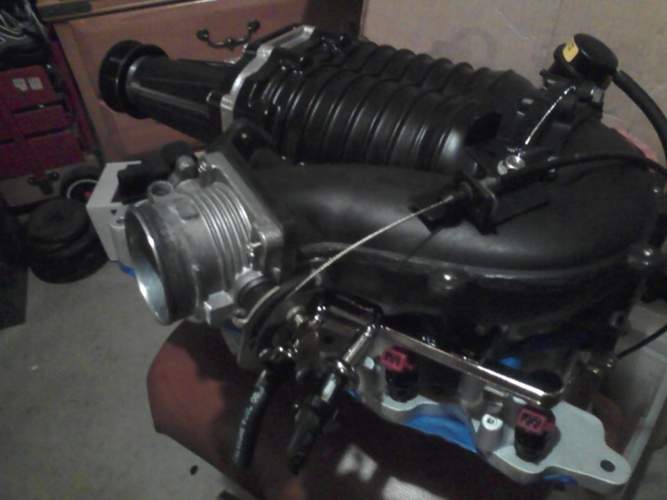 A test fit later on with flipped inlet.  

Note the now satin-black blower. The throttle body had to be flipped 180 to clear the new IAC location.  A small steel bracket was bent to secure the throttle cable. It is actually a saleen-included part bolted onto the small black bracket.  It is adjustable so I could take up the cable slack.


Also note the modified fuel rail. The two spring lock couplings were spares attached to test fitment - which was good because they would not have cleared the throttle linkage. The rail inlet/out were bent downward for more clearance. 


Getting to the injector connectors and COPs in this location (#5, #6, #7) is VERY difficult. The #6 plug is virtually inaccessible to replace, but is possible if it really had to be done.

I used all double platinum finewire Ford Lightning plugs (2 steps colder) so I wouldn't have to deal with them for a LONG time (rated for 100k service interval, so at least 50k and I hardly drive the car anyway)