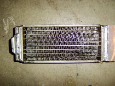 1996 Mercury Grand Marquis - A small aluminum heat exchanger or oil/fluid cooler with a finned core between two end tanks, resting on a dirty concrete floor. Weld beads…