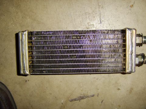 1996 Mercury Grand Marquis - A small heat exchanger or heater core lies on a dusty concrete floor, showing its finned aluminum core between two brass-colored end tanks…