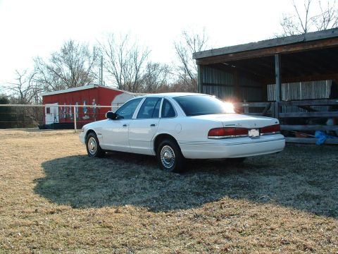 1995 Ford Crown Victoria - A white 1995 Ford Crown Victoria is parked on a grassy area next to a rustic wooden structure.