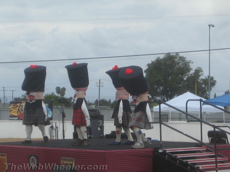 Four costumed mascots in kilts and tam hats perform on an outdoor stage at a Scottish festival