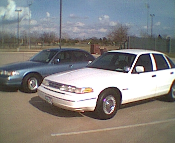 1995 Ford Crown Victoria - Two sedans, one white and one blue, are parked in a lot under a cloudy sky.