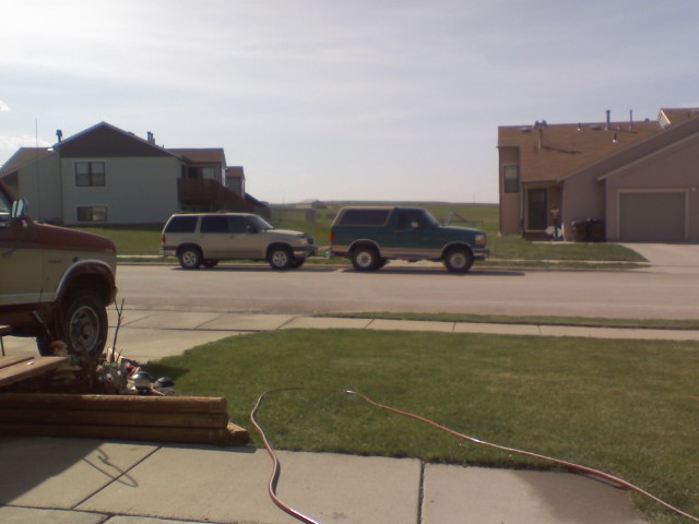 Green and white Ford Bronco parked curbside on a residential street, side view, silver SUV parked in front
