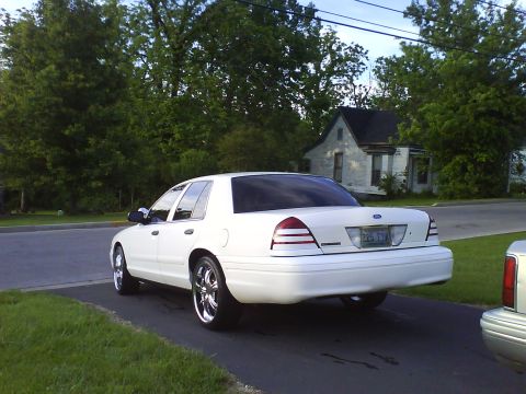 2003 Ford Crown Victoria - A white Ford Crown Victoria sedan is parked on a driveway with a house and trees in the background. The car has chrome rims and a license p…