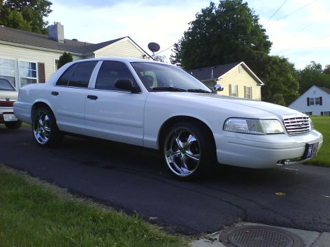 2003 Ford Crown Victoria - A white 2003 Ford Crown Victoria is parked on a paved driveway in front of a house. The car has tinted windows and chrome rims.