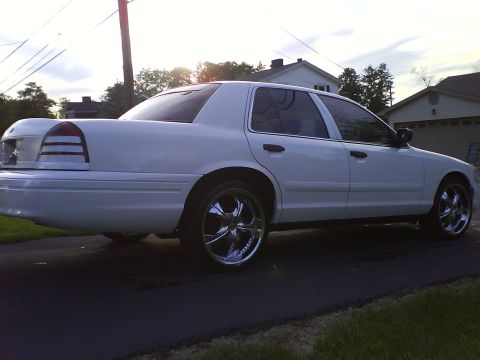 2003 Ford Crown Victoria - A white 2003 Ford Crown Victoria is shown from the rear quarter angle, parked on a driveway with chrome rims. The sun is setting behind the…