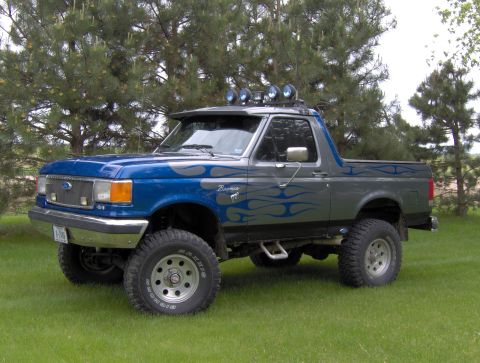 1987 Ford Bronco - A lifted, two-tone blue and gray 1987 Ford Bronco with blue flame graphics is parked on a grassy lawn in front of pine trees. The vehicle h…