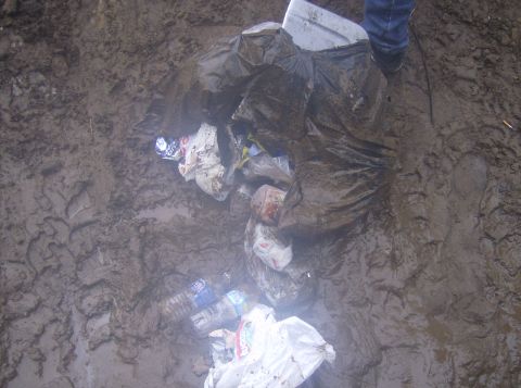 1986 Ford Bronco - A pile of trash bags and plastic bottles are strewn across muddy ground. A person's legs in jeans and boots are visible near the trash.