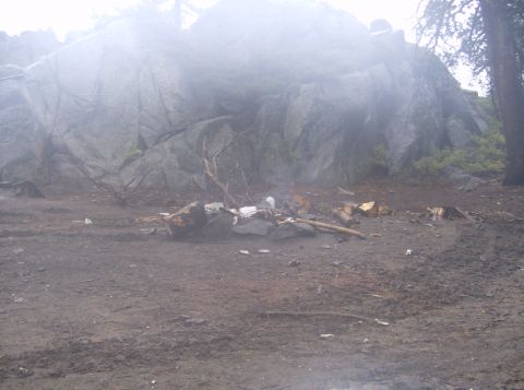1986 Ford Bronco - A campfire pit is filled with trash and debris in front of a large rock formation on a foggy day. The ground is muddy and littered with bra…