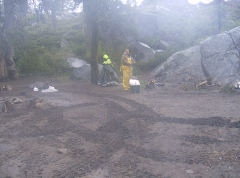 1986 Ford Bronco - Two people in rain gear stand in a muddy campsite with large rocks and trees. One person is near a tent, and the other is by a cooler.