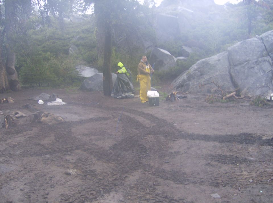 Two people in rain gear in a muddy campsite with rocks and trees.