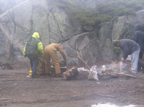 1986 Ford Bronco - Several people are gathered around a campfire in a rocky, misty outdoor setting. One person in a yellow rain jacket is standing, while othe…