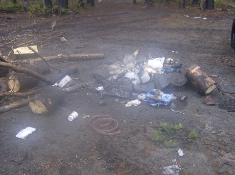 1986 Ford Bronco - This image depicts a messy campsite with a fire pit filled with trash, logs, and some ice. Debris is scattered around the muddy ground, inc…