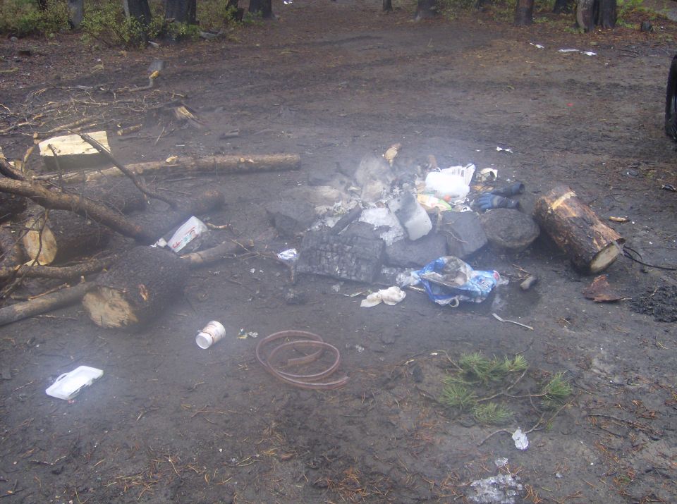 A messy campsite with a fire pit, trash, logs, and ice on muddy ground.