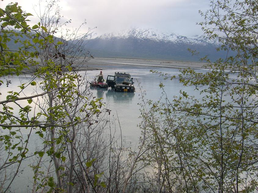 Off-road SUV and ATV partially submerged in a river with mountains in the background.