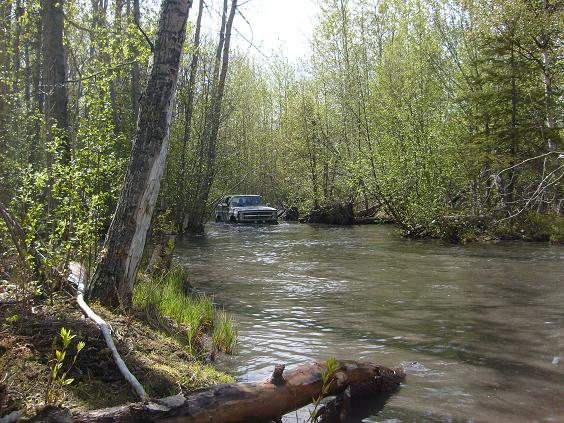 Dark Chevrolet SUV driving through a shallow river surrounded by trees.