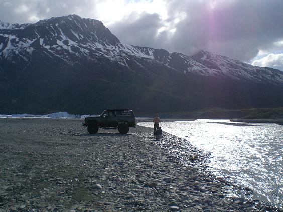 Dark SUV parked on a rocky shore by a river with mountains in the background.