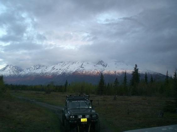 Dark SUV parked on dirt road with snow-capped mountains and forest in background.