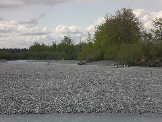 Dark vehicle on a rocky riverbed with trees and clouds.