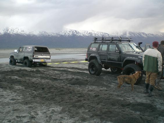Lifted dark SUV being towed by a black Jeep Cherokee on a sandy beach with mountains in the background.