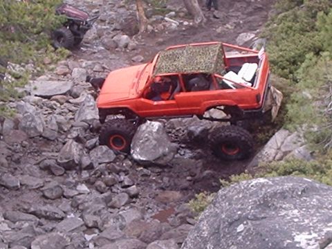 1986 Ford Bronco - An orange off-road vehicle with large tires navigates a rocky, muddy terrain. The vehicle has a camouflage-covered roof and appears to be m…