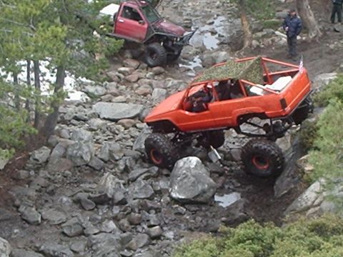1986 Ford Bronco - Two off-road modified trucks navigate a rocky, wet trail. The foreground vehicle is an orange SUV with a camouflage net over its roll cage,…