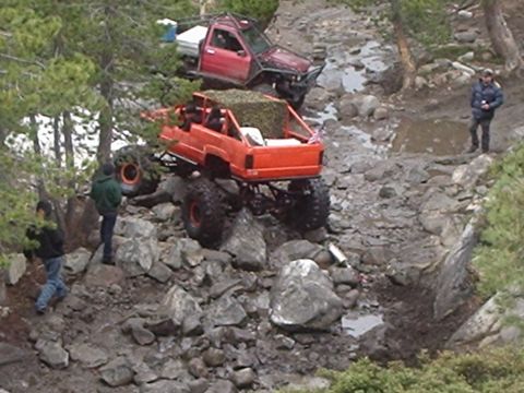 1986 Ford Bronco - Two off-road vehicles navigate a rocky, muddy terrain. An orange modified SUV with large tires is in the foreground, while a red modified S…
