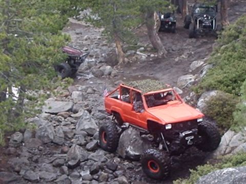 1986 Ford Bronco - An orange off-road SUV with large tires navigates a rocky, wooded trail. Another off-road vehicle is visible further up the trail.
