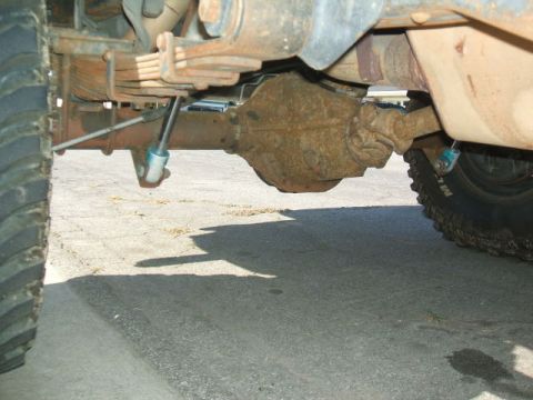 1988 Ford Bronco - Low under-vehicle view looking at the rear axle of a lifted truck, showing the surface-rusted differential housing, multi-leaf spring pack…