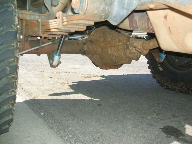 Underside view of a lifted truck's rusty rear axle, leaf springs and mud-terrain tires
