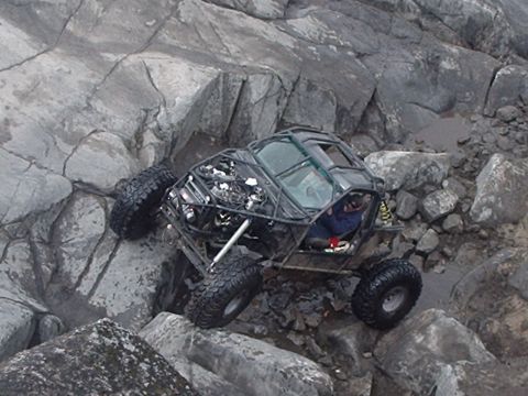 1986 Ford Bronco - A heavily modified off-road buggy with large tires navigates a rocky, wet terrain. The driver, wearing a blue jacket and cap, is visible in…