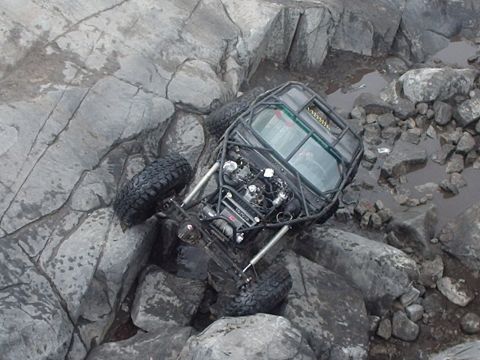 1986 Ford Bronco - A custom off-road buggy with a roll cage and exposed engine is navigating a rocky, wet terrain, tilted precariously on large boulders.