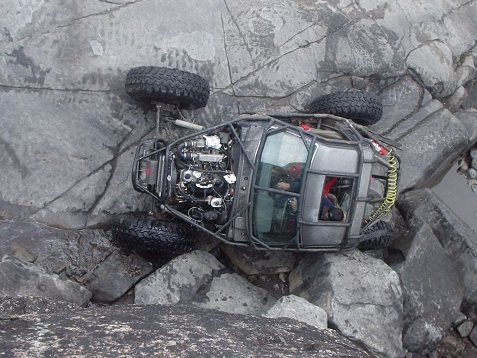 Off-road buggy tipped on its side on rocks, showing exposed engine and roll cage.