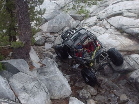 1986 Ford Bronco - A custom-built rock crawler is navigating a steep, rocky trail. The vehicle has a roll cage and large off-road tires, with its suspension a…