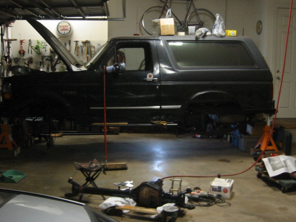 Dark Ford Bronco in a garage with hood up, on jack stands, with tools and parts on the floor.