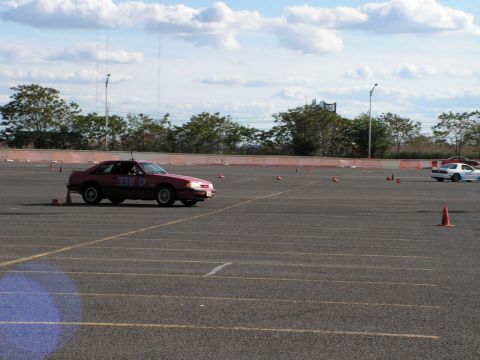 1990 Ford Mustang - A red coupe navigates an autocross course marked by orange cones under a partly cloudy sky.