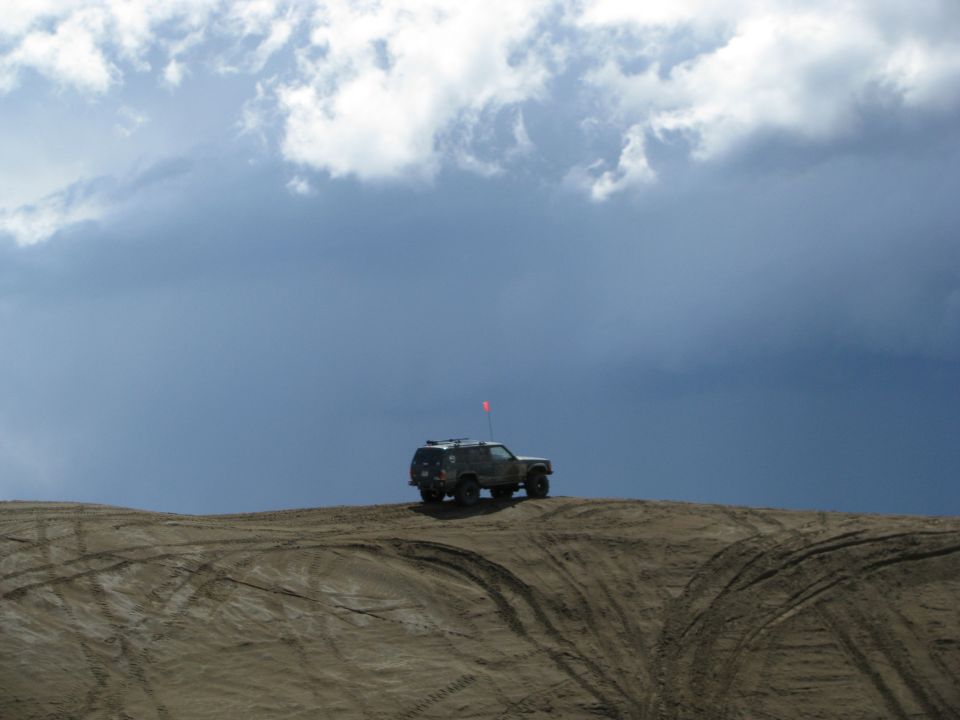 Lifted dark SUV with orange whip flag parked atop a sand dune under storm clouds