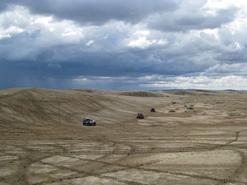 1994 Ford Bronco - Multiple off-road vehicles traverse sand dunes under a dramatic, cloudy sky.