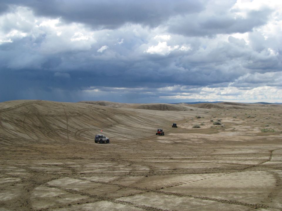 Dark lifted SUV, red sand rail and another rig on track-covered dunes under storm clouds