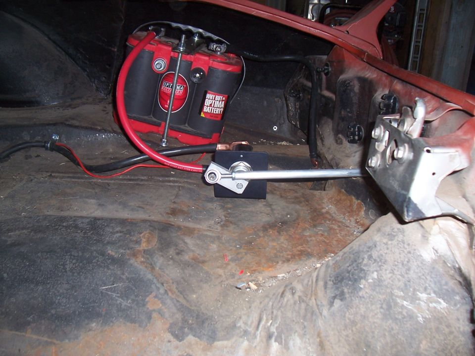 Car interior showing a red Optima battery and a silver shifter linkage on a rusty floor