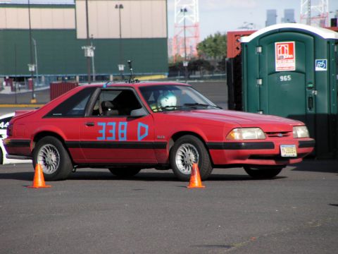 1990 Ford Mustang - A red 1990 Ford Mustang coupe, identified by its body style, is seen in a side profile with blue racing numbers on the door.