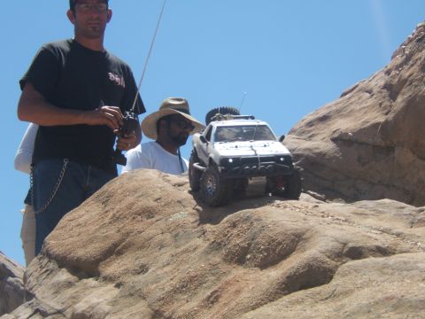 1988 Ford Bronco - Two people observe a remote-controlled off-road vehicle navigating a rocky landscape under a clear blue sky.
