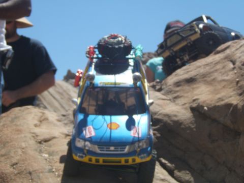 1988 Ford Bronco - A blue remote-controlled off-road vehicle navigates a rocky terrain, with another RC vehicle visible in the background.