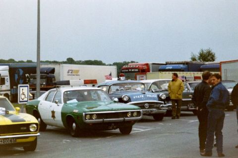 1991 Ford Crown Victoria - A police car with a star badge and light bar is parked in a lot with several other vehicles and people.