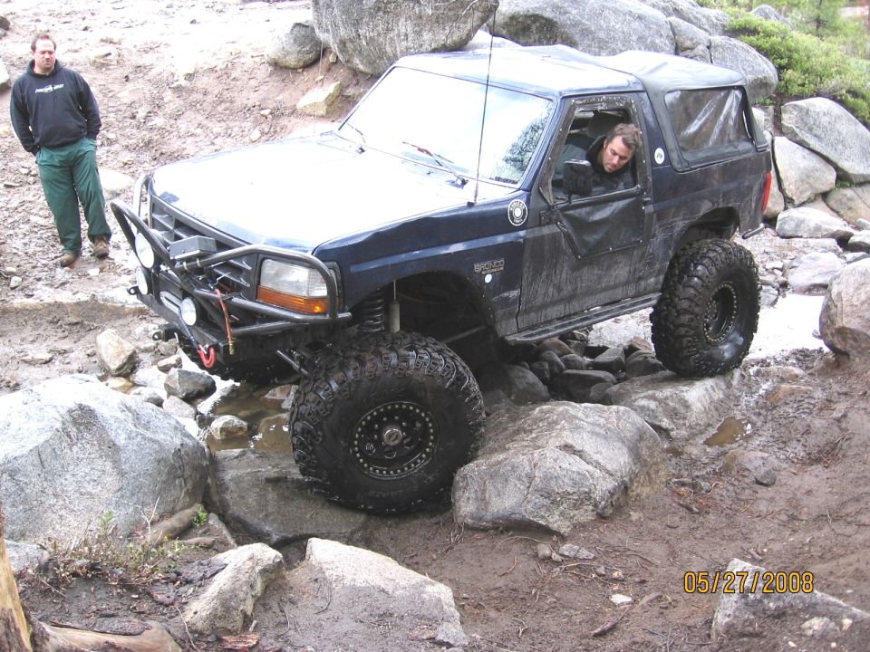 Lifted navy Ford Bronco rock-crawling over boulders in a creek with a front winch bumper