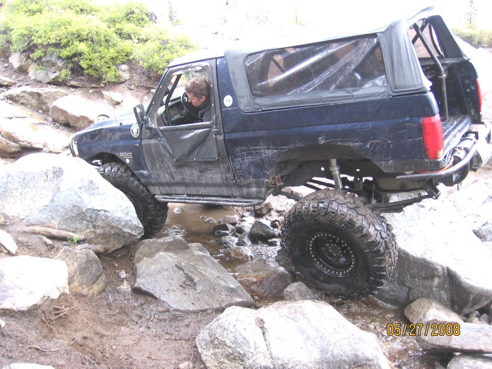 Doorless dark blue Ford Bronco on beadlock wheels crawling wet boulders on a rocky trail
